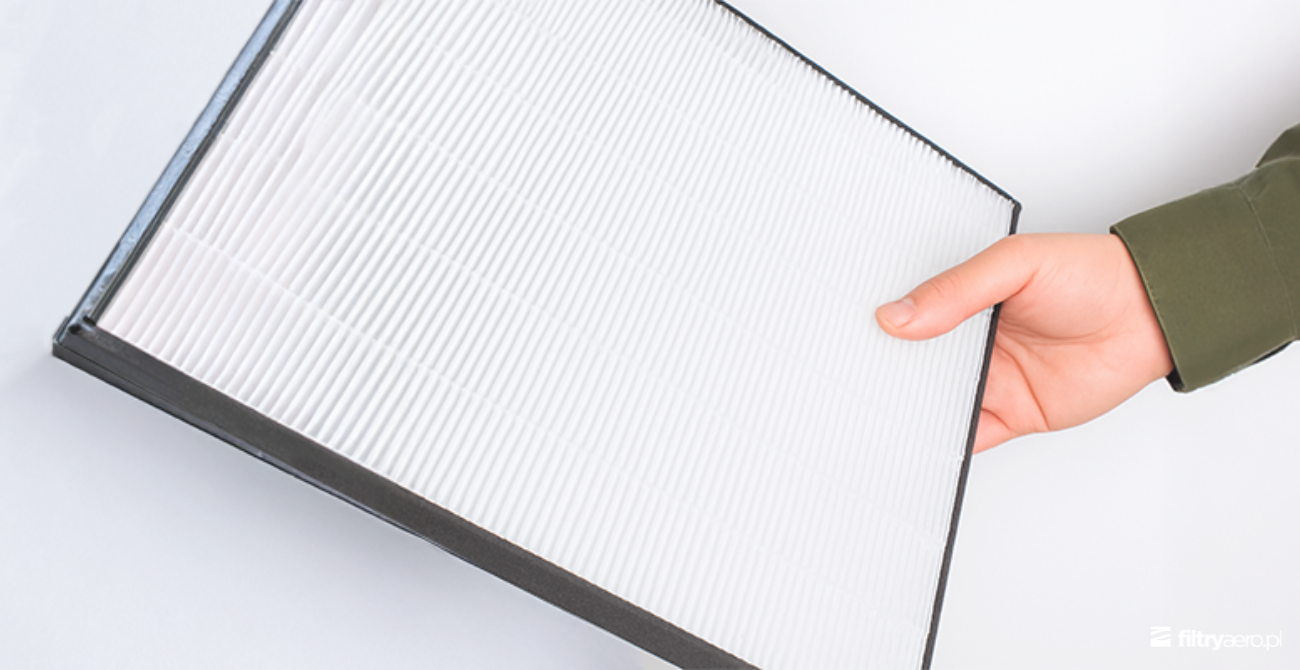 Dłoń trzymająca biały, gęsty filtr HEPA o strukturze harmonijkowej w czarnej ramce. Widoczna precyzyjna struktura materiału filtrującego.